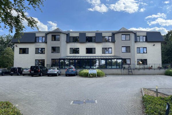 De volledige voorgevel en parking van woonzorgcentrum Beerzelhof, gezien vanaf de buitenkant. Het moderne gebouw wordt omringd door groene bossen, die bijdragen aan de rustige en natuurlijke uitstraling van de omgeving. De parkeerplaats voor het gebouw biedt voldoende ruimte voor bezoekers en bewoners. De groene natuur rondom het gebouw zorgt voor een vredige sfeer, ideaal voor ontspanning en rust.