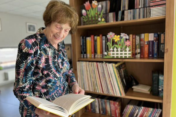 De ontspanningsruimte van woonzorgcentrum Aurora, waar bewoners boeken kunnen lenen en/of lezen. Op de achtergrond is een boekenkast te zien, gevuld met een gevarieerde collectie boeken. Een bewoner heeft een boek gepakt en is het rustig aan het doornemen.
