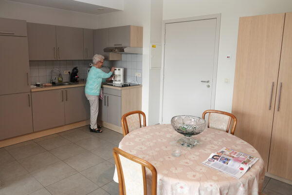 Een assistentiewoning in Aurora, waarbij de keuken en eetkamer zichtbaar zijn. De bewoner staat in de keuken en zet koffie met haar koffiezetapparaat. De eetkamer is ingericht met een ronde tafel en vier stoelen, en wordt opgefleurd door een grote glazen vaas als decoratie.