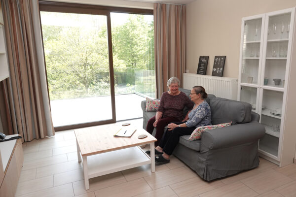 Twee bewoners zitten op een grijze sofa in de woonkamer van hun assistentiewoning, die is ingericht met een vitrine en een bijzettafel. Via een schuifdeur is het terras toegankelijk, dat is voorzien van glazen leuningen.